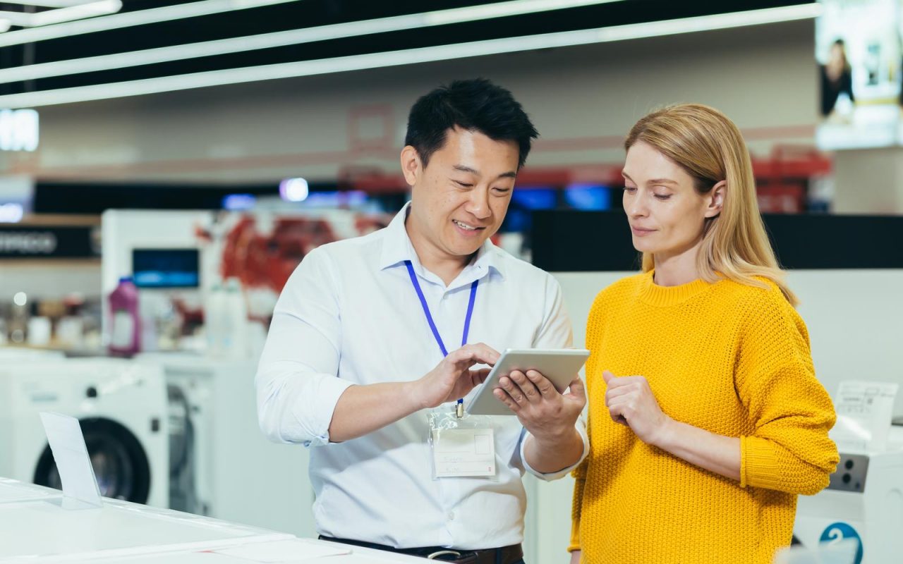 Asian consultant salesman in electronics and household appliances store, selling a working machine to a woman, recommending and approving the choice